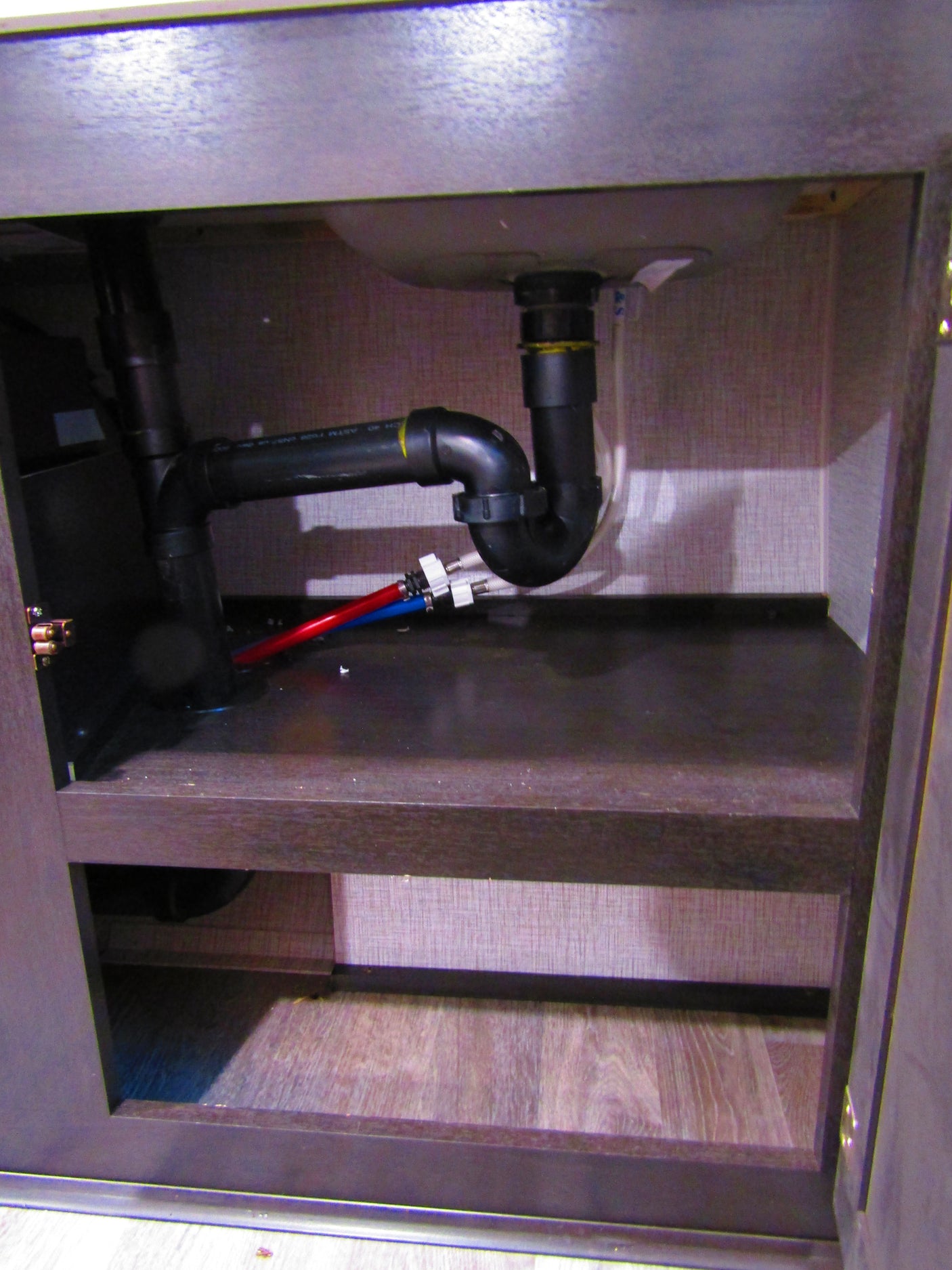 Inside an **RV rental** under-sink cabinet, revealing plumbing for the sink drain, P-trap, and red/blue water lines.