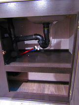 Inside an **RV rental** under-sink cabinet, revealing plumbing for the sink drain, P-trap, and red/blue water lines.