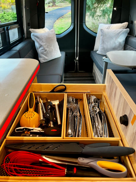 An RV rental's organized kitchen drawer: utensils, silverware, and tools for meal prep, with cozy interior seating and views.