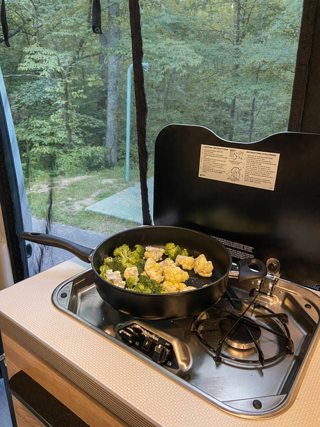 Cooking broccoli and cauliflower on a two-burner stove inside an RV rental, with a view of trees through a screen.