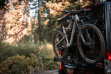 Dark RV rental with two mountain bikes on a rear rack, parked on a dirt road in a sunlit forest.