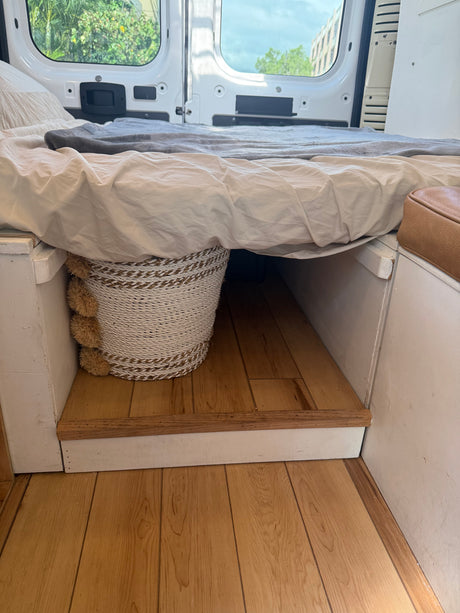 Comfortable bed setup in an RV rental, featuring under-bed storage with a woven basket. Open rear doors reveal outdoor scenery.