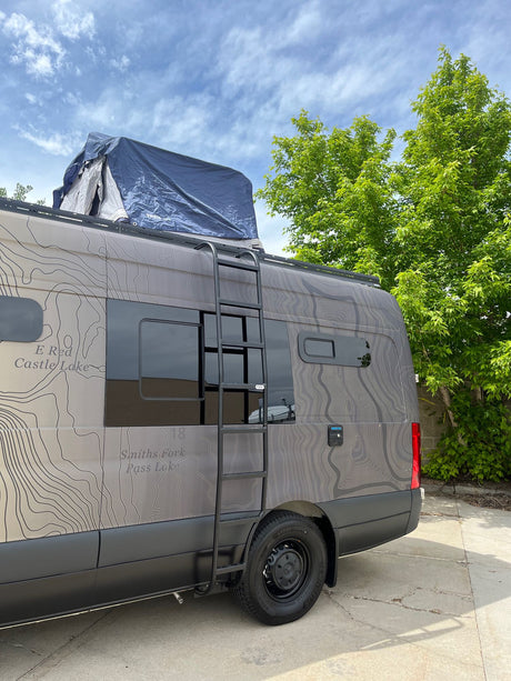 This custom rv rental camper van, adorned with a topographical map design, features a ladder and rooftop tent, ideal for adventure travel.