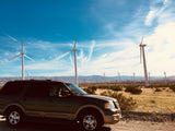 A dark SUV at a desert wind farm under blue skies. Ideal for road trips! Think RV rental for your next adventure.