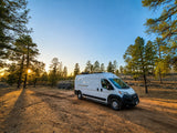 This white RV rental van is parked on a sunlit dirt road, surrounded by pine trees at sunset, ideal for nature camping.
