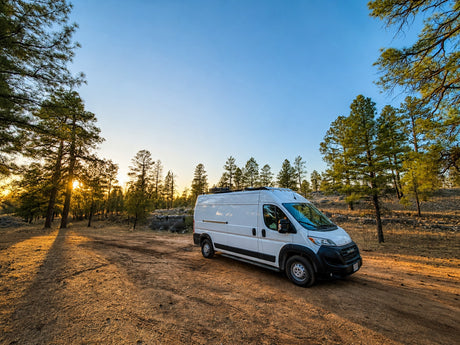 This white RV rental van is parked on a sunlit dirt road, surrounded by pine trees at sunset, ideal for nature camping.