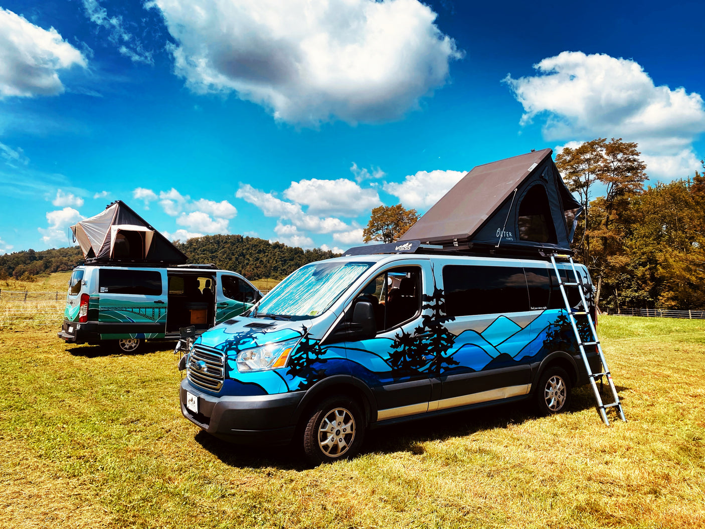 Two customized RV rental camper vans with rooftop tents in a scenic field under a blue sky.