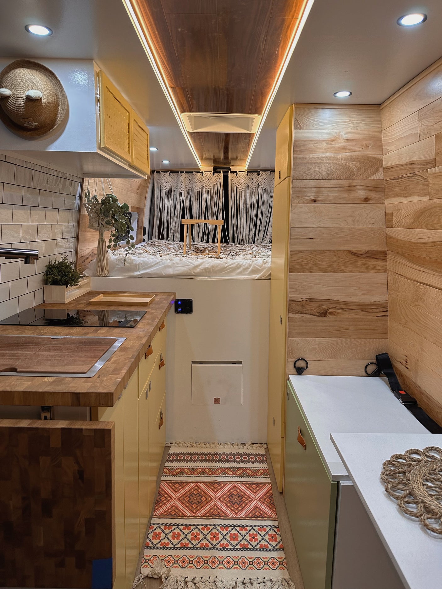 Cozy rv rental interior: compact kitchen, elevated bed with macrame, warm wood walls & cabinets. Functional and inviting.