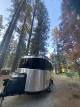 A silver rv rental Airstream is parked in a forest campsite, surrounded by tall pine trees. Ideal for nature getaways.
