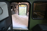 Inside an RV rental: a compact sleeping area with a polka dot comforter. The open rear hatch reveals a grassy outdoor view.