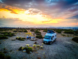 Scenic RV rental camping at sunset. A camper van, picnic table, and people are set up in a vast landscape.