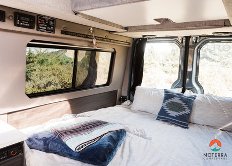 Cozy campervan RV rental interior showing a comfortable bed, window with green views, and control panels.