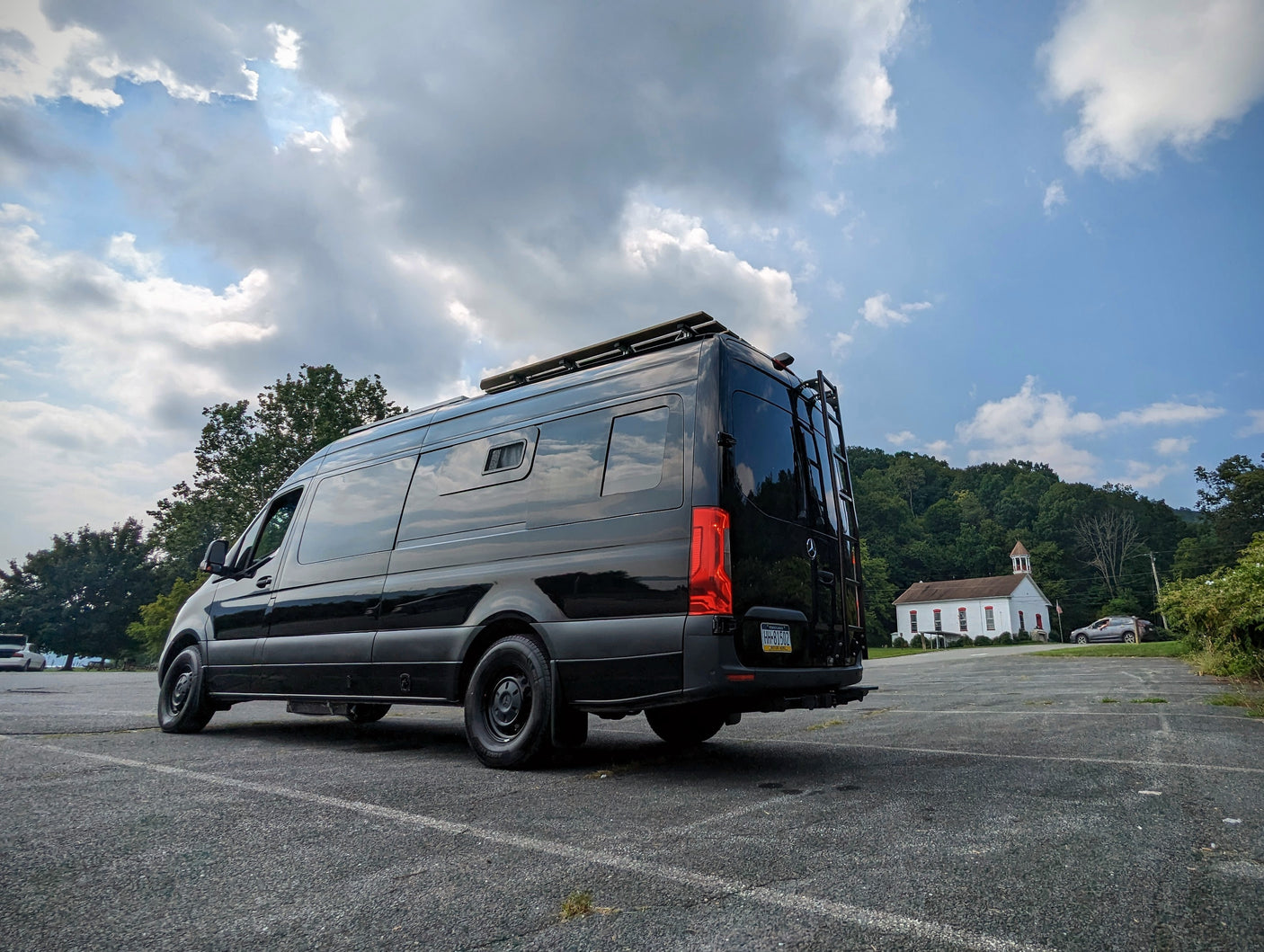 This black camper van, perfect for RV rental, is parked in a scenic lot with a charming church and green hills in the background.