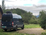 Experience adventure! This dark camper van RV rental is parked in a picturesque mountain landscape with forest & cabin nearby.