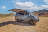 Adventure-ready gray camper van with extended awning, parked on a dirt lot amidst mountains. Ideal rv rental for outdoor exploration.