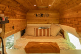 Inviting RV rental interior: wood-paneled walls, comfortable bed, pillows, and a "WELCOME" sign. Recessed lights illuminate the space.