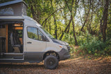Your next adventure starts with this RV rental! Light-colored camper van, pop-top, off-road ready, parked in a forest.