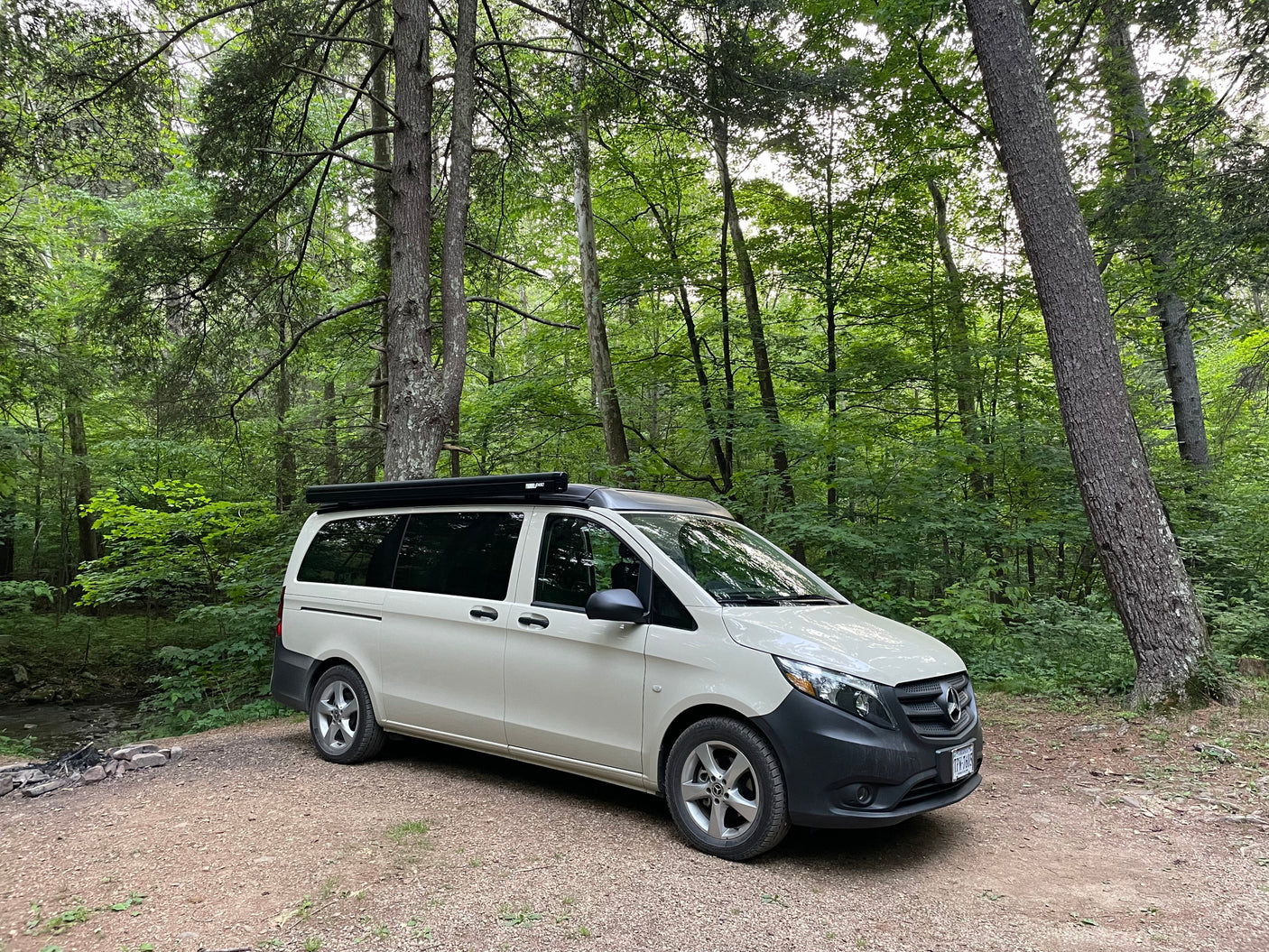 White Mercedes-Benz camper van parked by a fire pit in a lush forest. Perfect RV rental for camping & nature exploration.