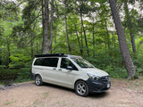 White Mercedes-Benz camper van parked by a fire pit in a lush forest. Perfect RV rental for camping & nature exploration.