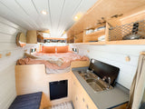 Inside this RV rental: a comfy bed, compact kitchen with stove/sink, and fridge. Bright, modern wood and white paneling.