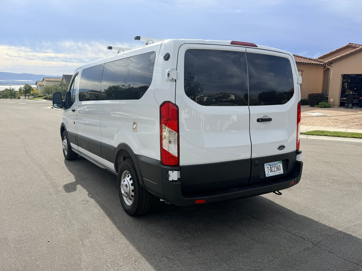 White Ford Transit rv rental van with roof racks & Montana plate, parked on a street with houses and hills in background.