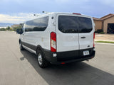 White Ford Transit rv rental van with roof racks & Montana plate, parked on a street with houses and hills in background.