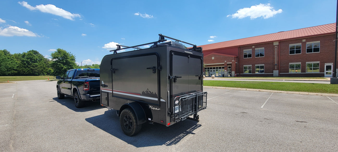 This Flyer RV rental trailer, hitched to a pickup truck, is parked in a lot near a brick building under a sunny sky.