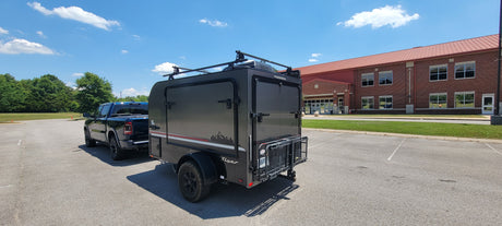 This Flyer RV rental trailer, hitched to a pickup truck, is parked in a lot near a brick building under a sunny sky.