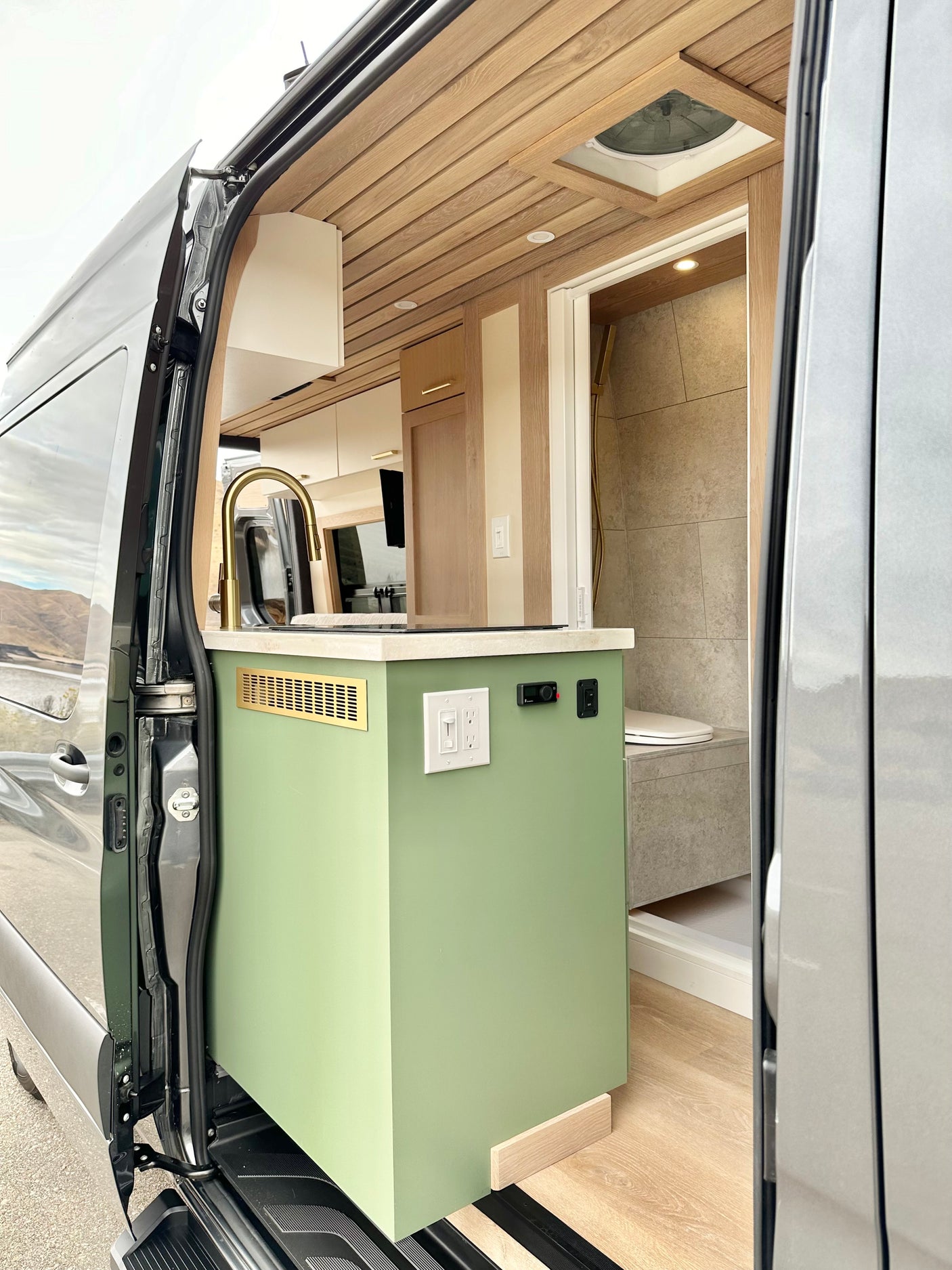 View inside an **rv rental**, showing a sage green kitchen with gold faucet & a compact shower/toilet area.