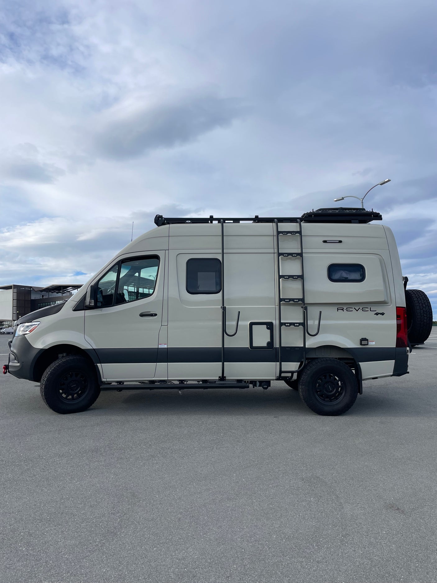 This beige Sprinter RV rental, equipped for off-road adventure with a roof rack and ladder, is parked on asphalt.