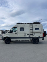This beige Sprinter RV rental, equipped for off-road adventure with a roof rack and ladder, is parked on asphalt.