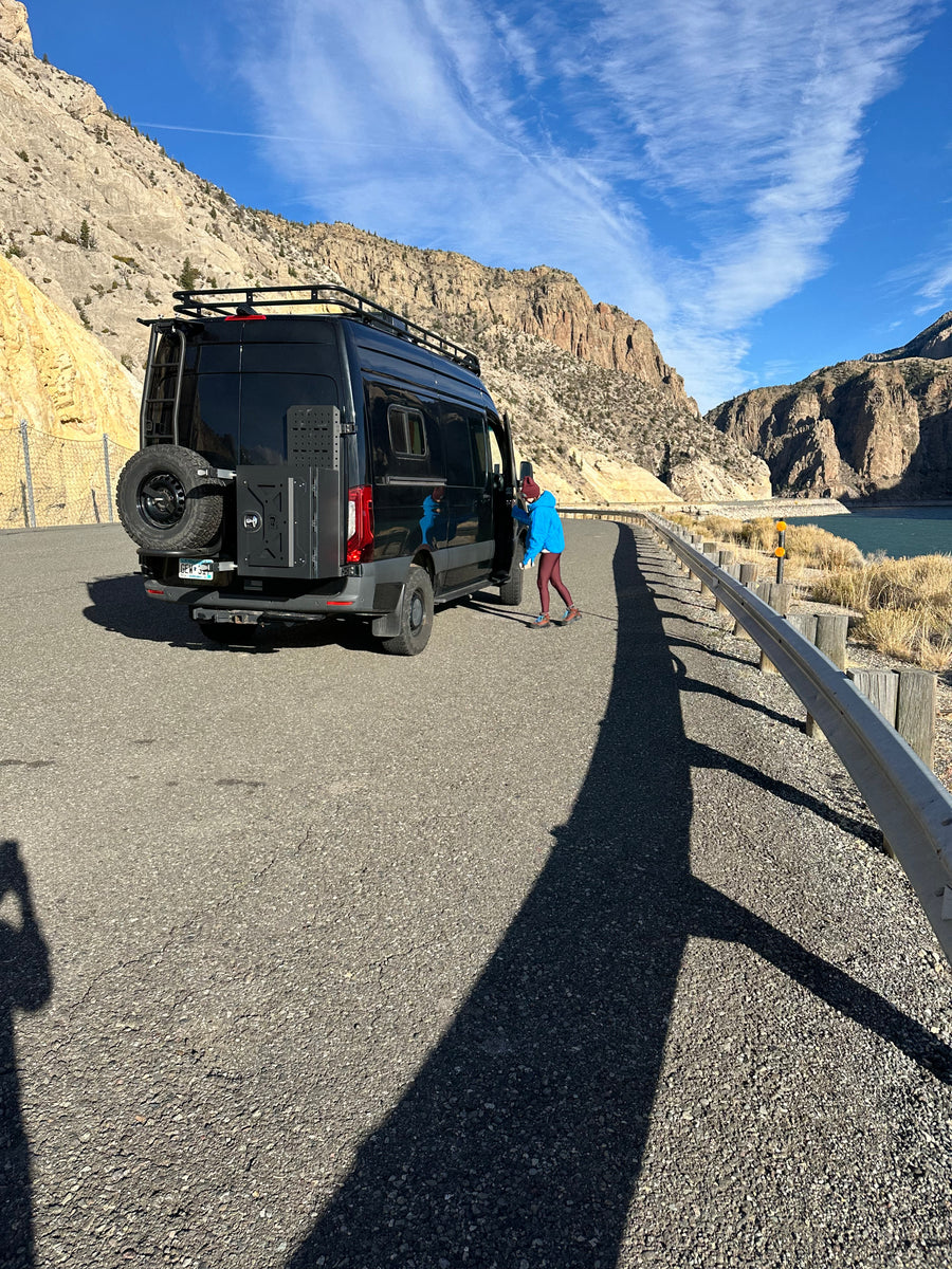 A black rv rental camper van is parked on a mountain road beside a lake, with a person standing by its door under blue skies.