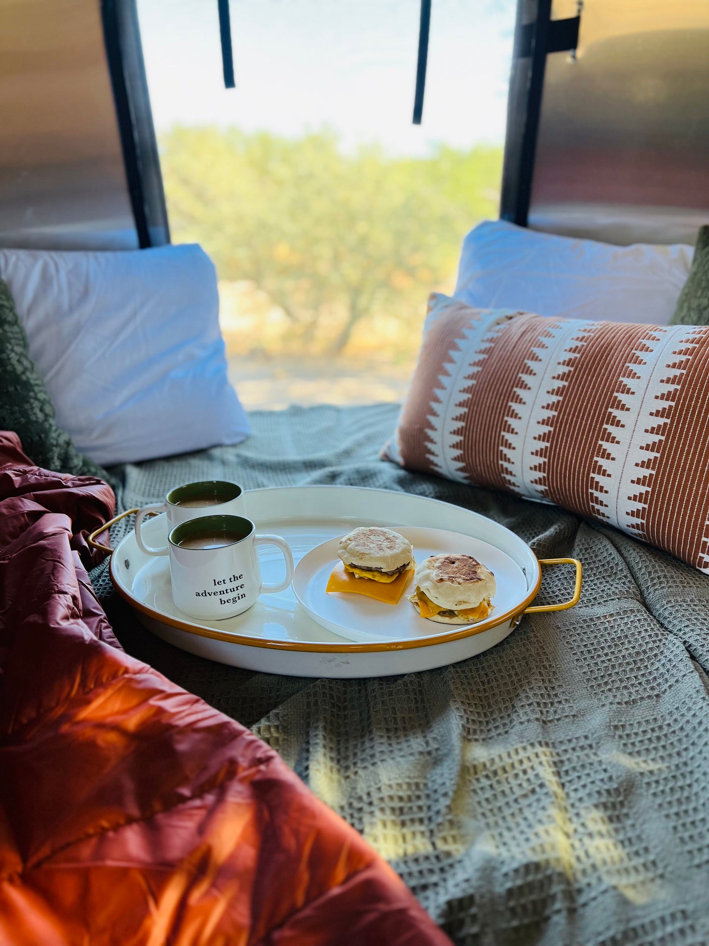 Breakfast in an RV rental: two mugs, two sandwiches on a tray with comfy pillows & outdoor views. Let the adventure begin!