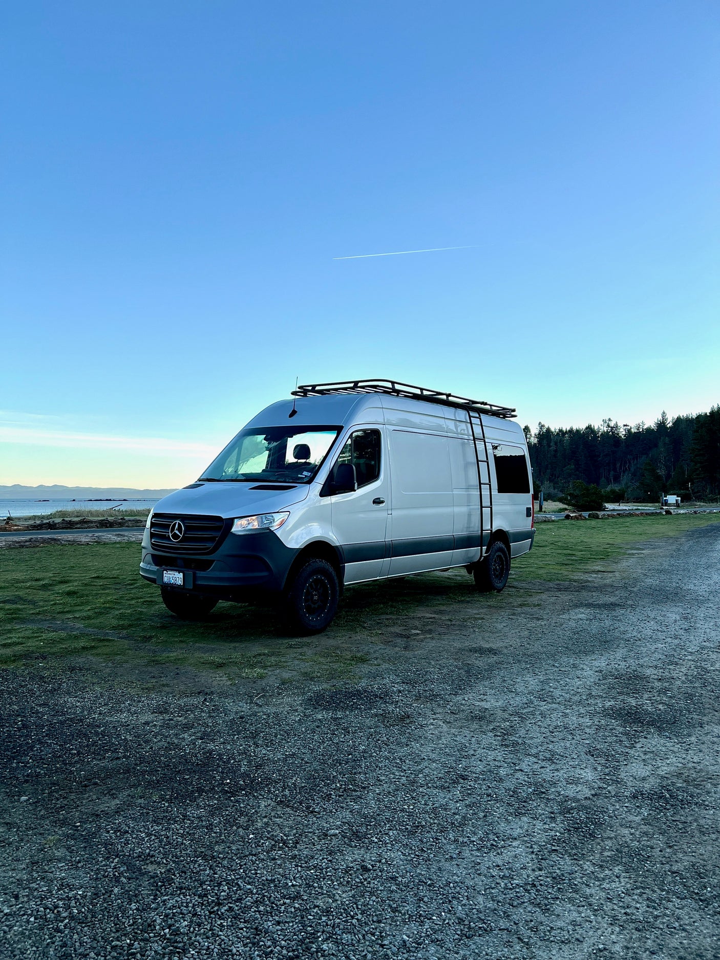 Experience van life with this silver Mercedes Sprinter rv rental parked beside a serene coastal landscape and forest.