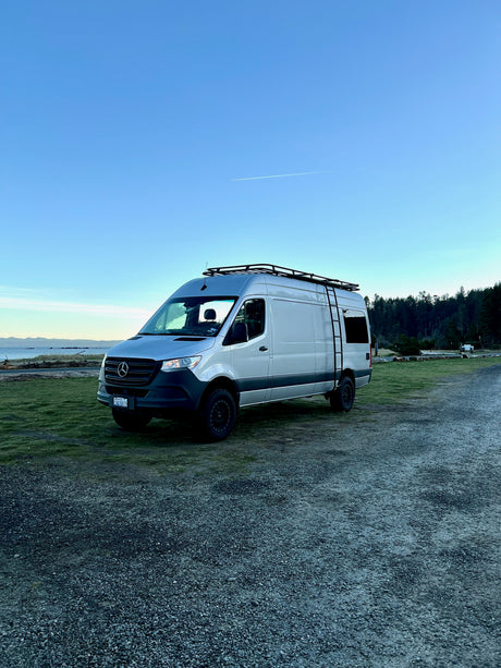 Experience van life with this silver Mercedes Sprinter rv rental parked beside a serene coastal landscape and forest.