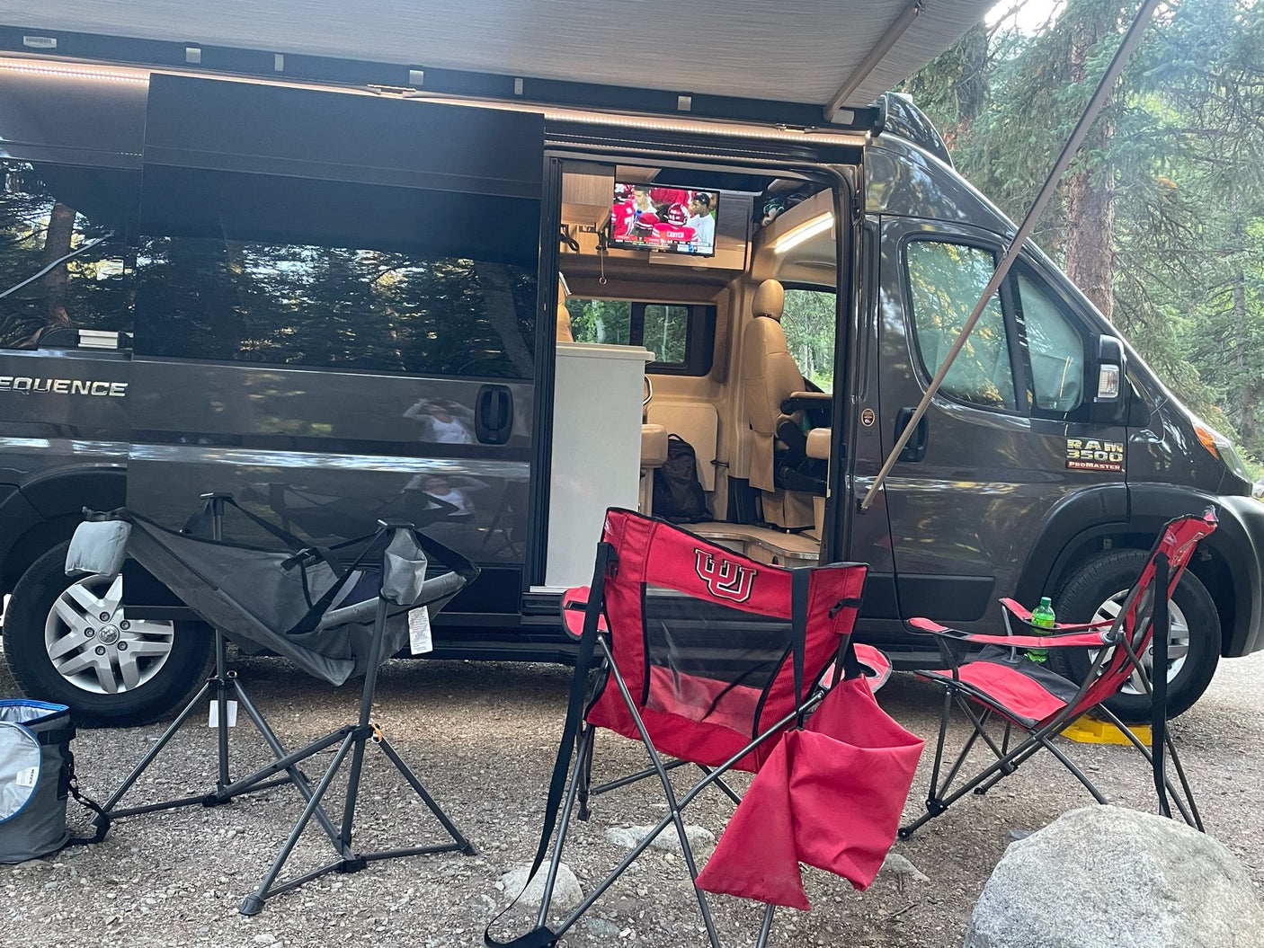 RV rental camper van with awning out at a gravel campsite. Red chairs (Utah Utes!) are set up, and a TV plays inside.