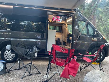 RV rental camper van with awning out at a gravel campsite. Red chairs (Utah Utes!) are set up, and a TV plays inside.