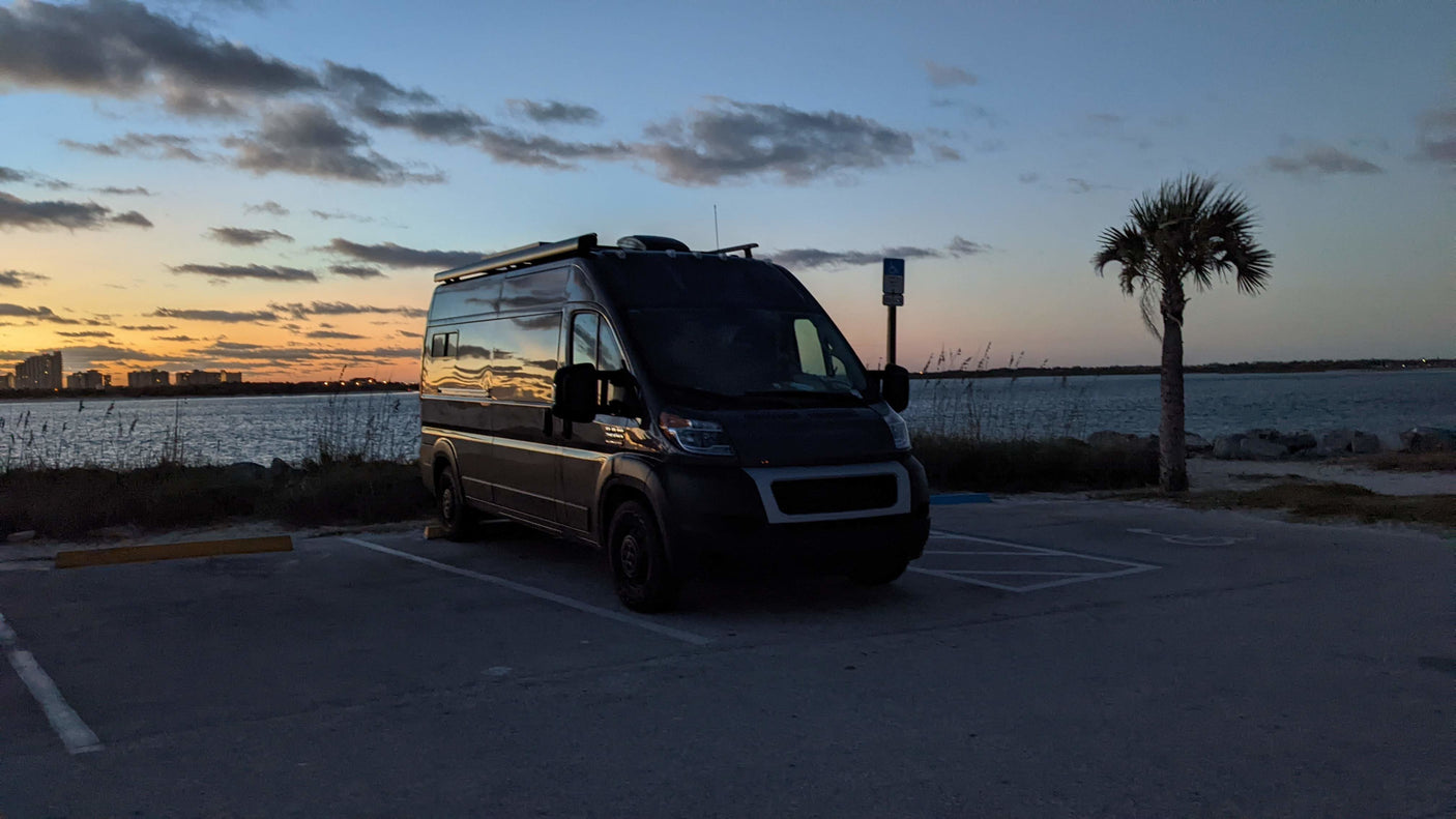 Adventure awaits! This dark RV rental camper van, perfect for travel, reflects the sunset by a waterfront with a city skyline.