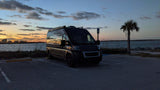 Adventure awaits! This dark RV rental camper van, perfect for travel, reflects the sunset by a waterfront with a city skyline.