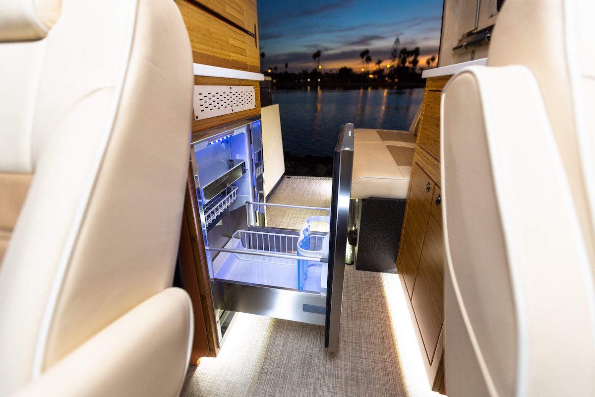  and control panels. Spacious mobile living space.|Enjoy beach sunsets in an RV rental! Camper van interior with kitchen view to girl surfing at golden hour.|RV rental rear: bench seat above utility box with power plug & Quick Charge. Floor shows hose & lighting.|This RV rental's interior reveals a sleek kitchen — photo 7