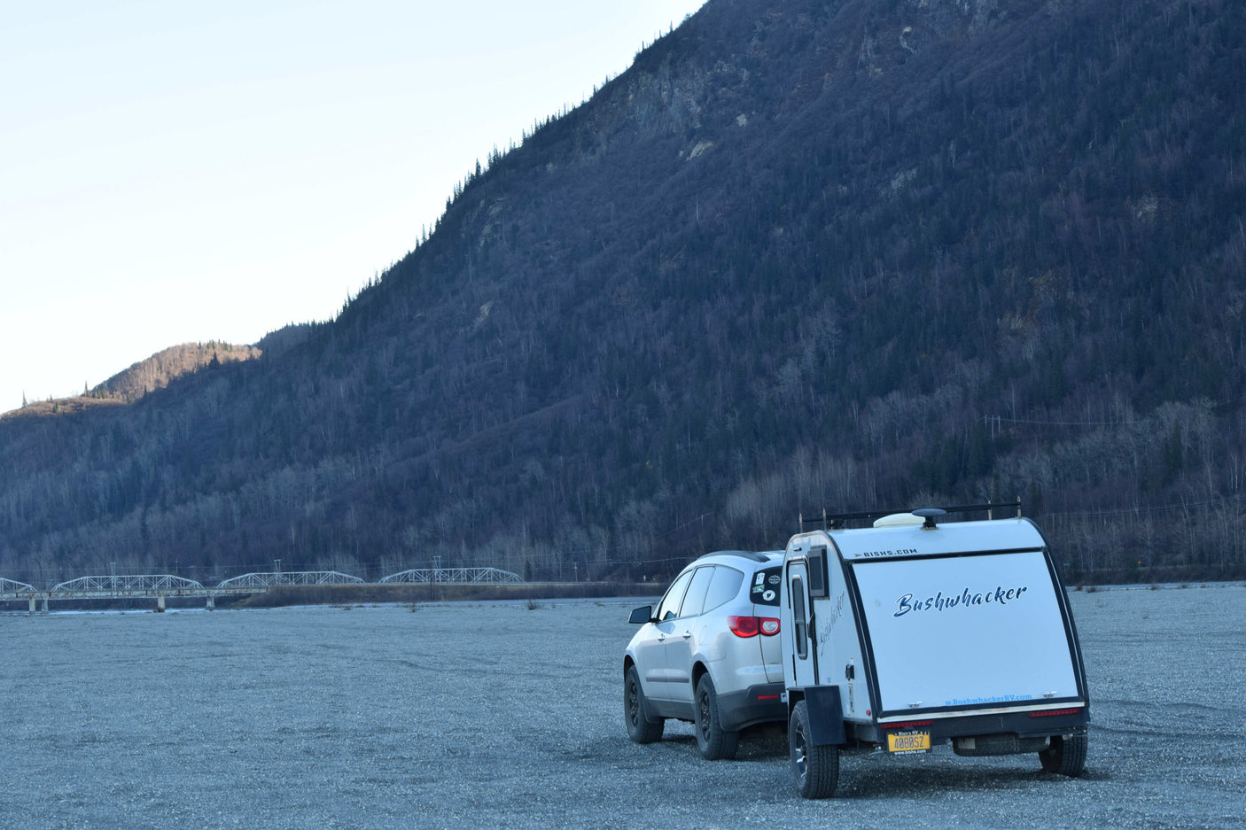 White SUV and Bushwhacker teardrop RV rental parked on gravel, with a mountain and bridge in the background.
