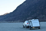 White SUV and Bushwhacker teardrop RV rental parked on gravel, with a mountain and bridge in the background.