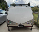 Rear view of a compact RV rental teardrop trailer, adorned with travel stickers, a roof rack, and deployed stability jacks.