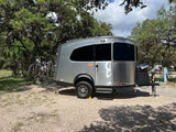A sleek, silver travel trailer, perfect for an rv rental, is set up at a shaded campsite with bikes on the back.