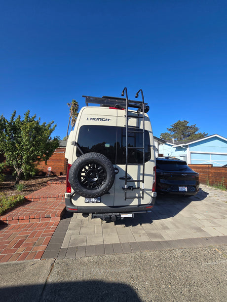 Rear view of a beige camper van, an RV rental, with a roof rack and a rear-mounted spare tire, on a sunny day.
