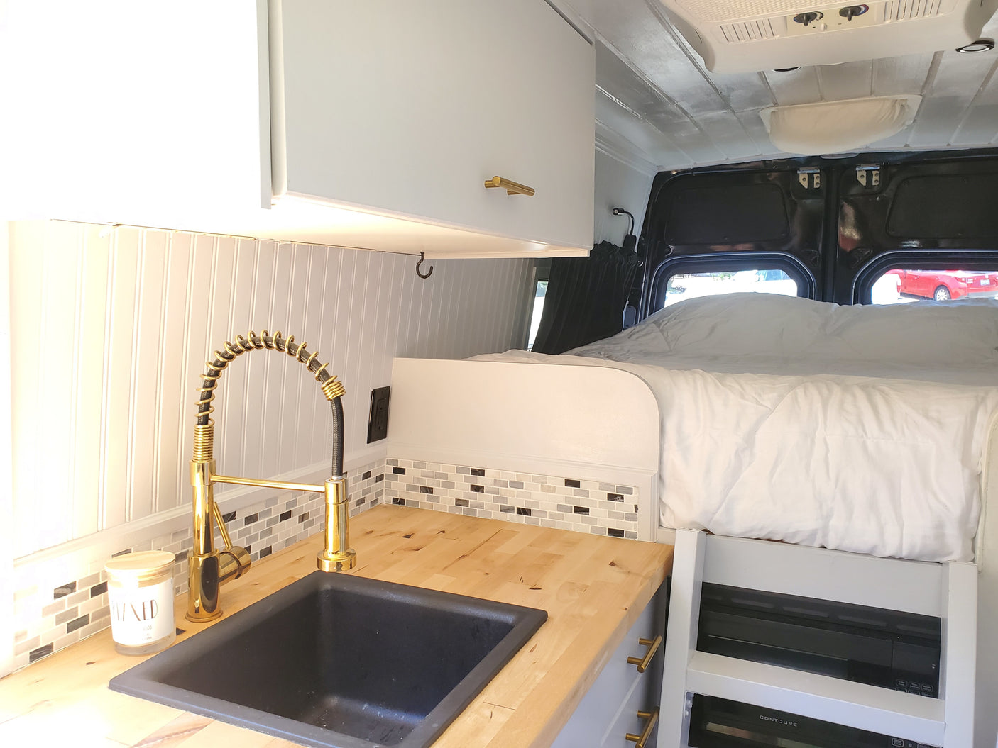 RV rental kitchenette: wood counter, black sink, gold faucet, mosaic backsplash. Features a cozy bed with white linens.