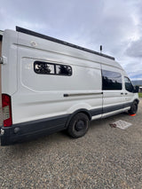Side view of a white Ford Transit camper van parked on gravel, suggesting an RV rental ready for adventure.