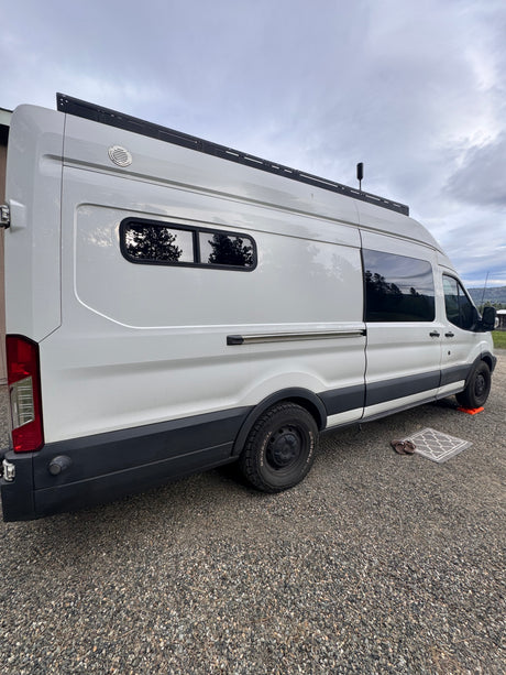 Side view of a white Ford Transit camper van parked on gravel, suggesting an RV rental ready for adventure.