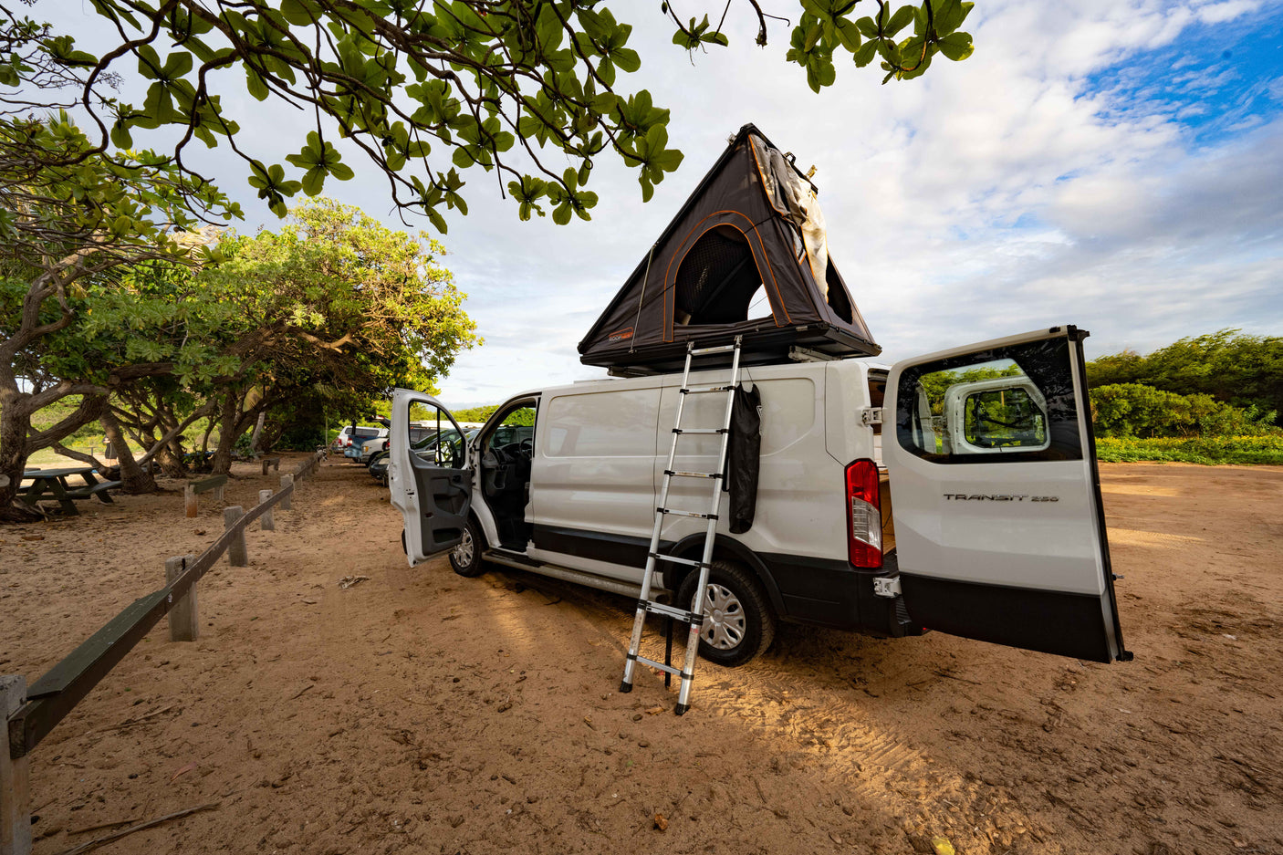 White Ford Transit rv rental with a Roofnest rooftop tent & ladder, doors open, parked on sand in a tree-lined campsite. Perfect for travel!