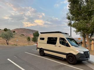 Tan Mercedes Sprinter RV rental van parked on a road, with scenic hills & trees under cloudy skies. Perfect for adventure.